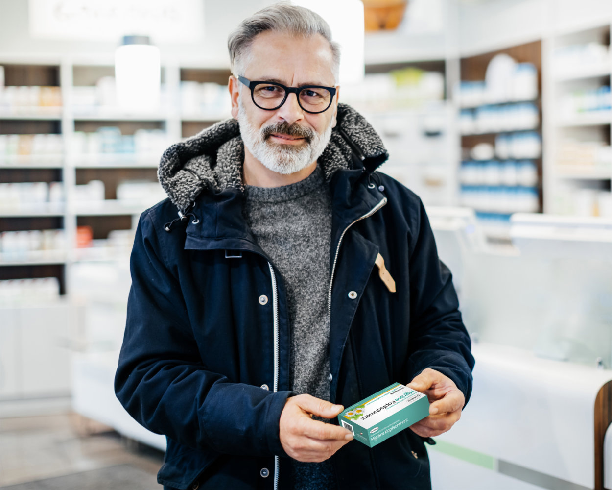 Mann mit grauem Bart und schwarzen Brillen steht in der Apotheke und hält eine Packung Migräne Tablettenin der Hand